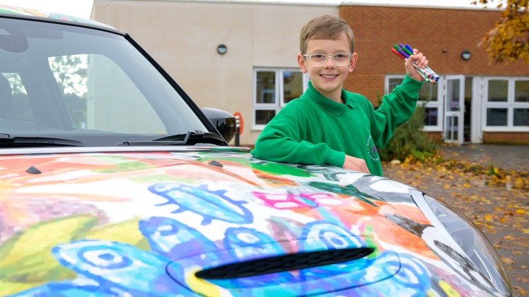 Close-up of the colorfully foiled MINI Cooper hood. Behind it, smiling Oliver Gorrod holding up Crayola pencils.