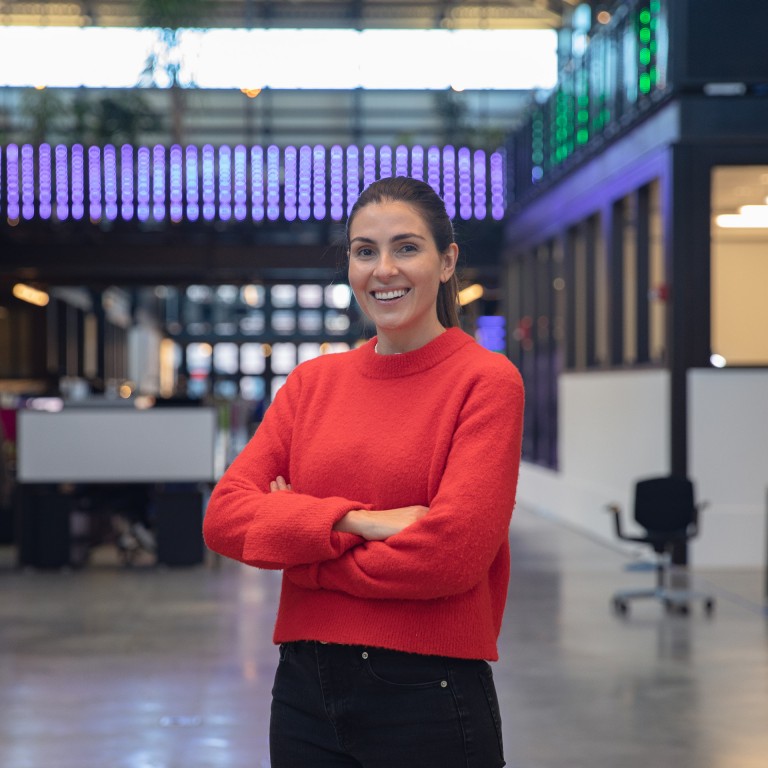 ¾ portrait of Miriam Roure, smiling at the camera. She stands in the co-working space of Newlab, where URBAN-X moved to in 2021.   