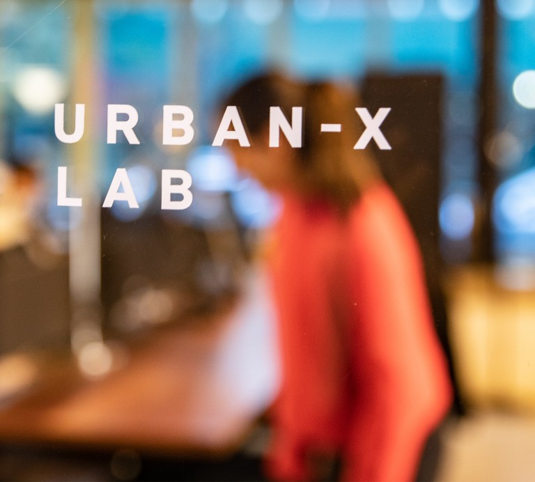 Image of the blurred silhouette of Miriam Roure, who is behind a glassdoor with the imprint “URBAN-X Lab”. 