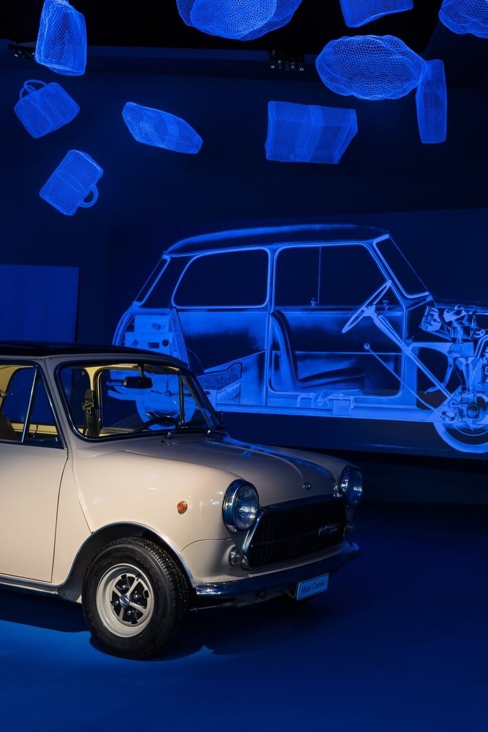 A white vintage MINI parked in front of a black light poster of its inner workings.