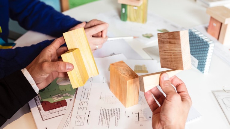 Building blocks are held in hands and placed on top of building plans.