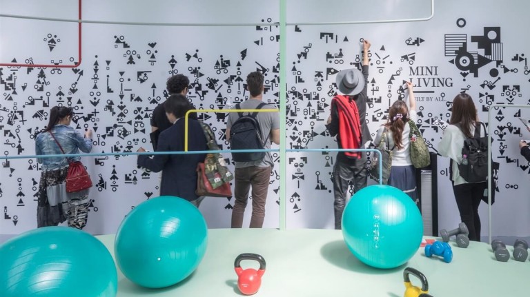 Young men and women explore the MINI LIVING exhibit at Salone del Mobile 2018. Colourful exercise equipment is in the foreground.