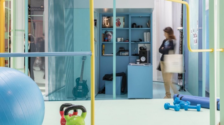 Woman stands in a tiny blue study containing a blue desk and stool as well as a black guitar. Exercise equipment is visible in the foreground.