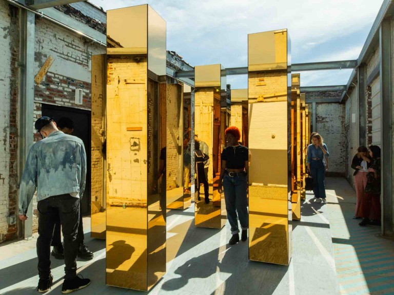 People walk among tall, yellow mirrored columns surrounded by white brick buildings.	