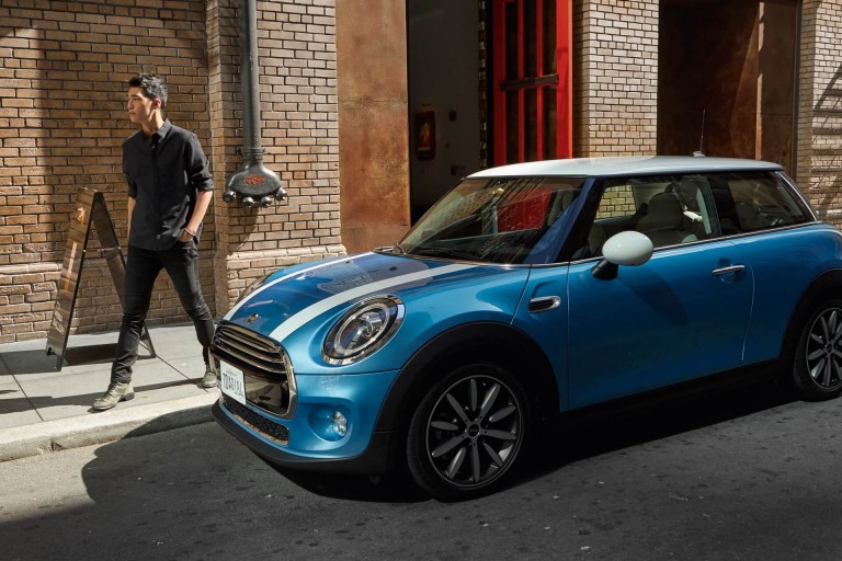 Young guy standing in the sun next to his Digital Blue MINI 3-Door with white roof and bonnet stripes.