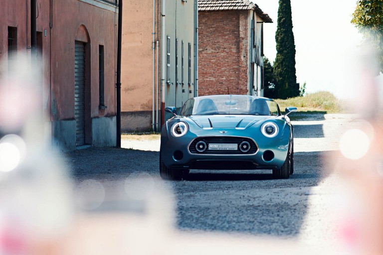 MINI Superleggera driving down a street with picturesque buildings, front view.