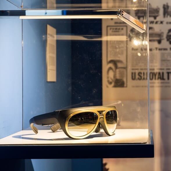 Gold racing glasses with a built-in camera are displayed in a glass case.