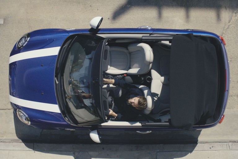 Overhead view of man driving MINI Convertible along a coastal road in the sunshine with top folded down.