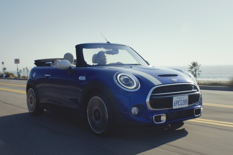 Front/side view of a MINI Convertible driving along a coastal road in the sunshine with top folded down.