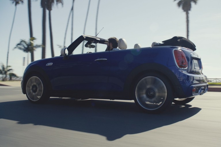 Side view of man driving a MINI Convertible along a coastal road in the sunshine with top folded down.