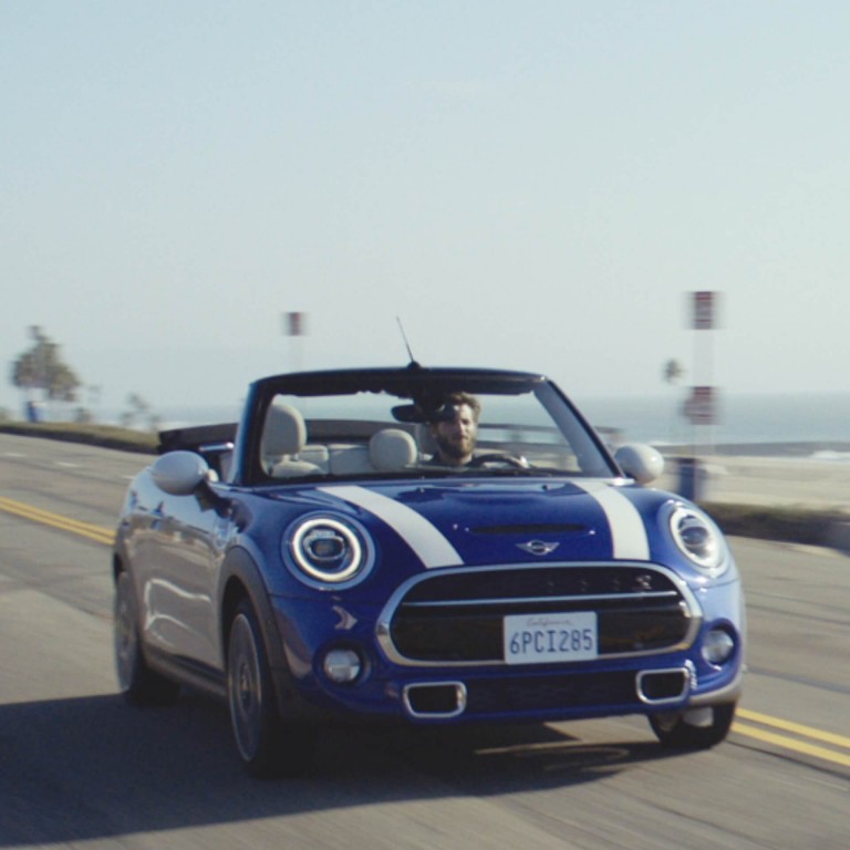 Front view of a MINI Convertible driving along a coastal road in the sunshine with top folded down.