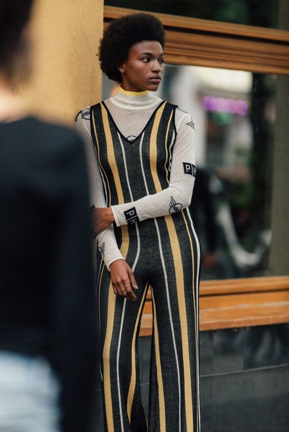 A woman, leaning on a yellow wall wears a MINI turtle neck pullover and a striped overall by PH5.