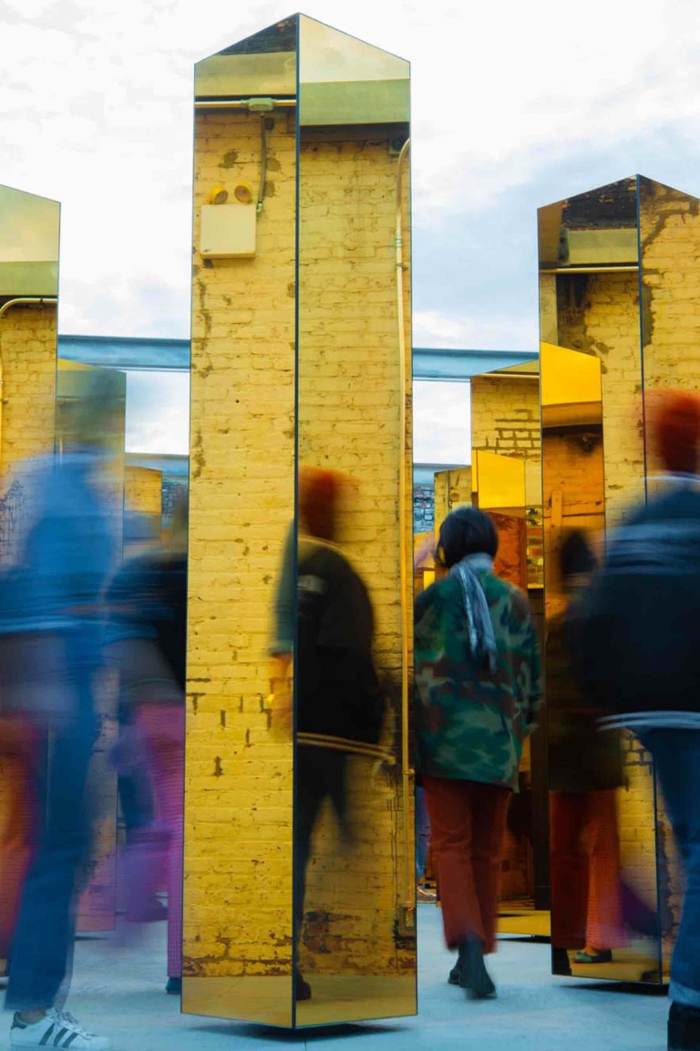 Image floue de visiteurs passant entre les colonnes en miroirs dorés de l’installation.