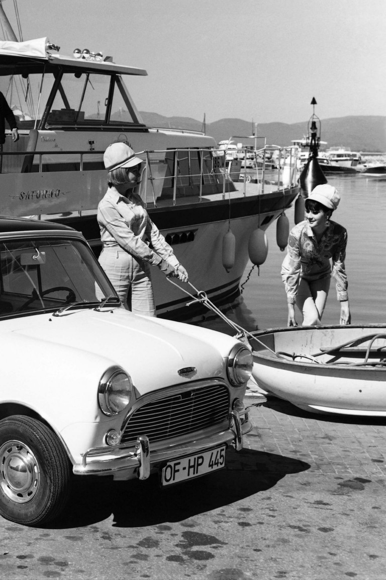  Morris Mini-Minor parked by the water's edge at a marina and two women prepare to board a dinghy nearby.