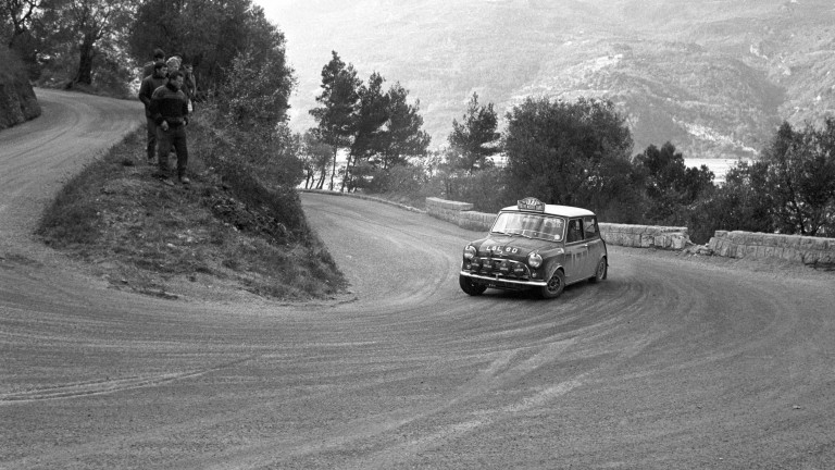 Spectators watch the BMC Works Mini Cooper S fly around a turn at the 1965 Monte Carlo Rally with trees and mountains in the background.