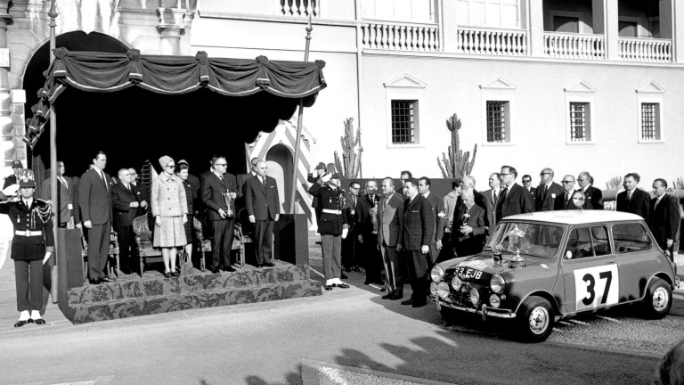 The BMC Works Mini Cooper S wins the 1965 Monte Carlo Rally. Trophies are awarded in front of a historical building.