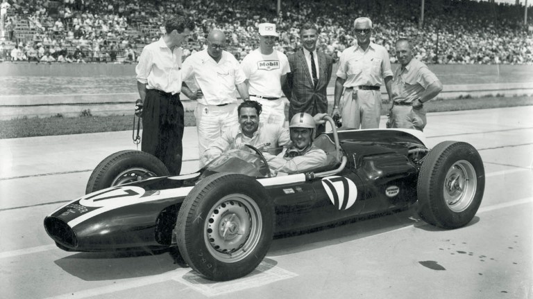 Cooper takes the rear engine revolution to Indianapolis in 1961. Driver sits in race car while Cooper kneels beside and others look on, full stands in the background.