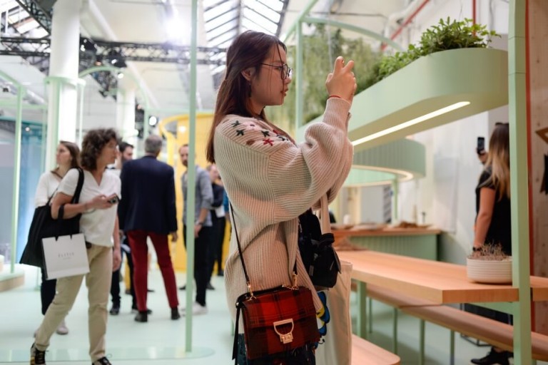 A young woman photographs part of the MINI LIVING exhibition while standing in the communal area. Other visitors are visible in the background.