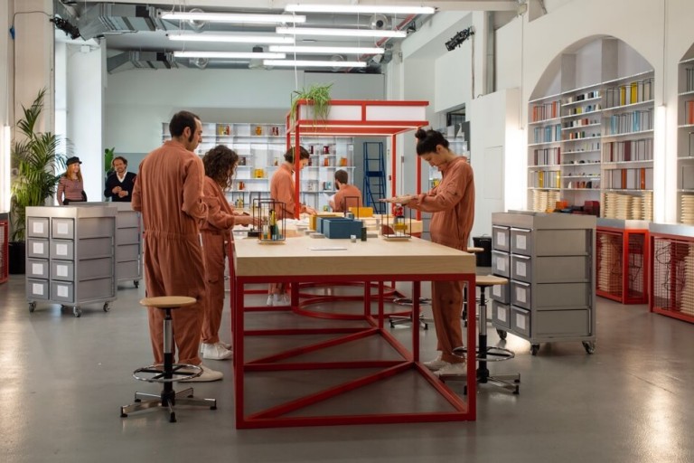 Men and women in peach coloured jumpsuits make models for new living spaces while gathered around a long table.