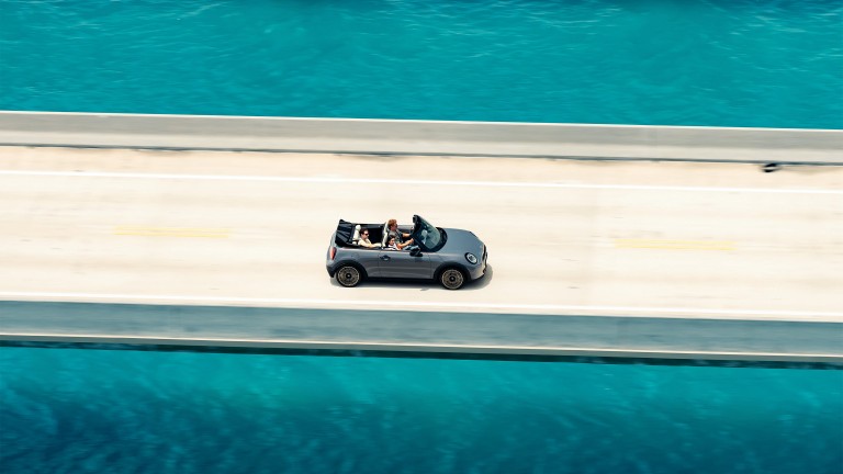 Overhead shot of the new MINI Cooper Convertible with an open top and three occupants, being driven down a road that has turquoise blue water at either side.