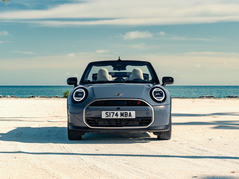 Head-on view of the new MINI Cooper Convertible, top down, with blue skies and the beach in the background.