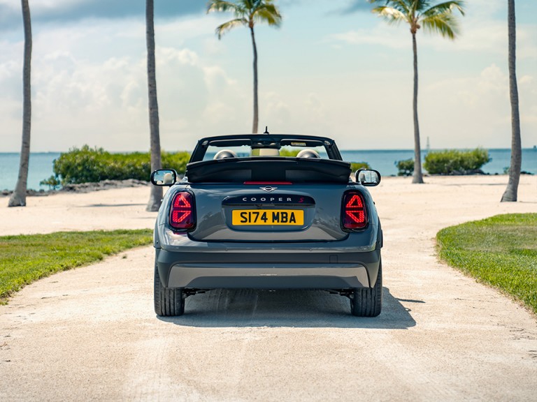 Pictured from the back, the new MINI Cooper Convertible with the top down and palm trees and the sea in the background.