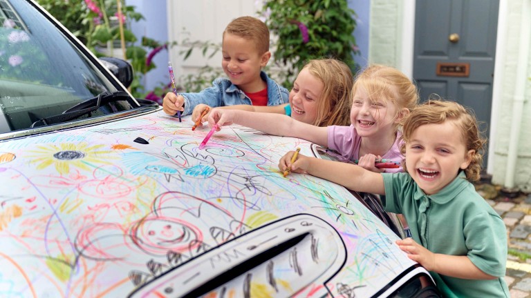 Picture of four laughing children who are painting the white hood of a MINI Cooper with Crayola pencils.