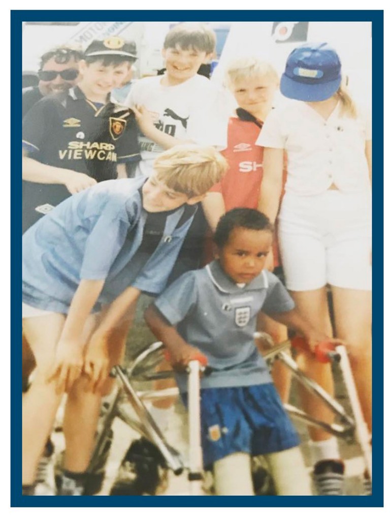 Nic as a little child surrounded by kids. He holds on to a walking aid and looks into the camera. 