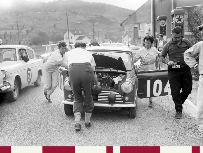 Black and white image of Pat Moss and Ann Wisdom at the Monte Carlo Rallye in 1962. They are slowly driving a Mini Cooper through a crowd of men. The plate number 737 ABL is placed prominently and across the bonnet. Also a red white graphic element is included below the image.