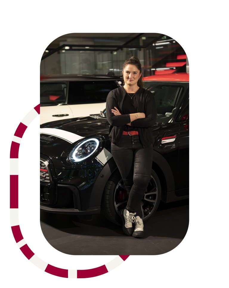 Image of the MINI color and material designer Morgane Bavagnoli, who developed the Pat Moss Edition. She´s standing in front of the Midnight Black John Cooper Work model from the Pat Moss Edition. Outside the image, there´s a graphic element in red and white.