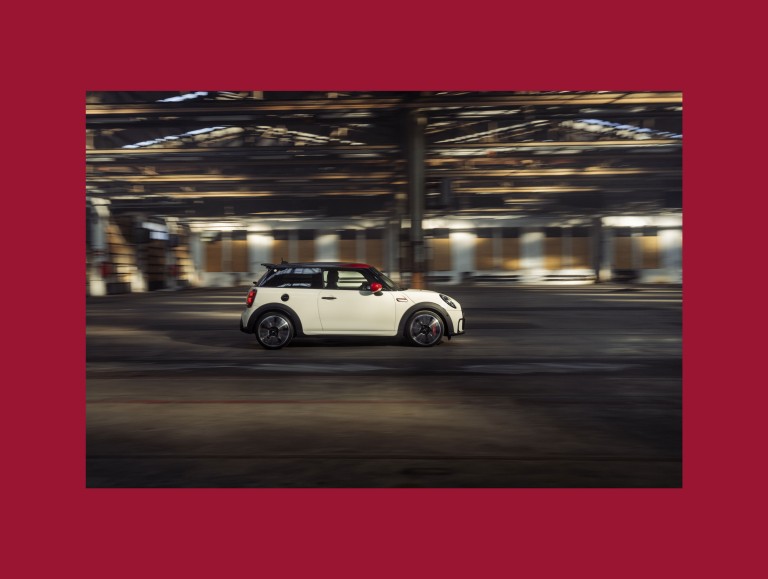 On a red background the following is placed: A fully side view of a driving John Cooper Works in the color Pepper White, from the Pat Moss Edition. 