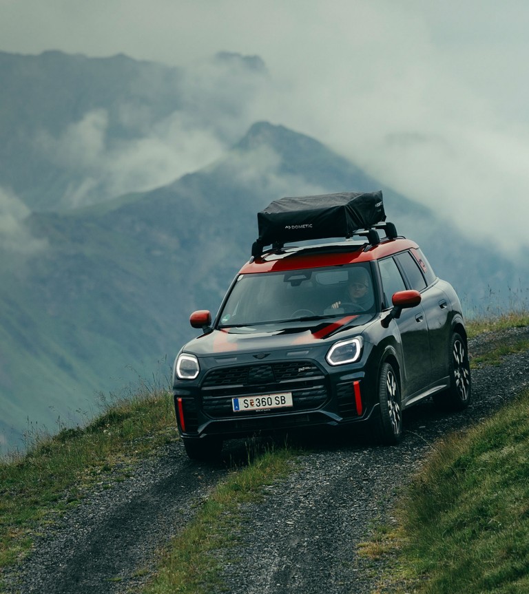 The new MINI John Cooper Works Countryman ALL4 with roof rack pictured driving around a corner on an unpaved mountain road, with a steep drop-off and misty mountain views in the background.