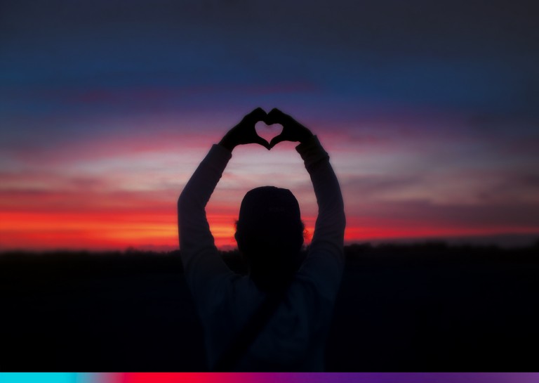 Image of a person at sunset. The person holds up her hands and makes a heart shape in the spirit if BIG LOVE UNITED. 