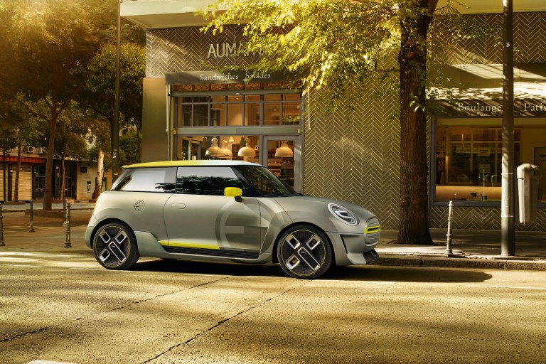 A silver and yellow MINI Electric Concept car, shown from the side, is parked in front of a retro-looking shop.