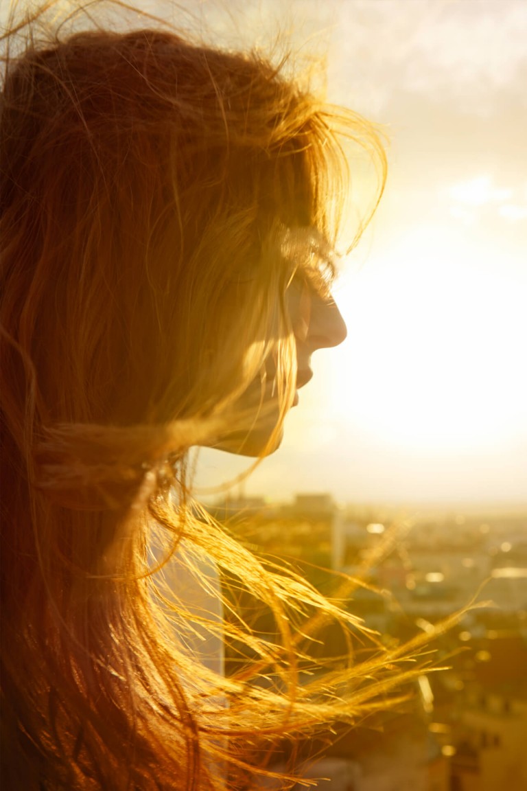 Young woman with long, flowing hair looks out over a city, sun shining in the background.