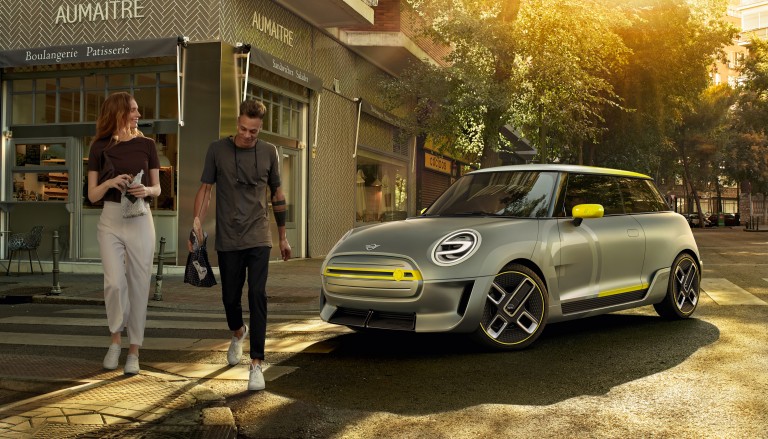 Stylish young woman and young man walk away from a shop and past a silver and yellow MINI Electric Concept car.
